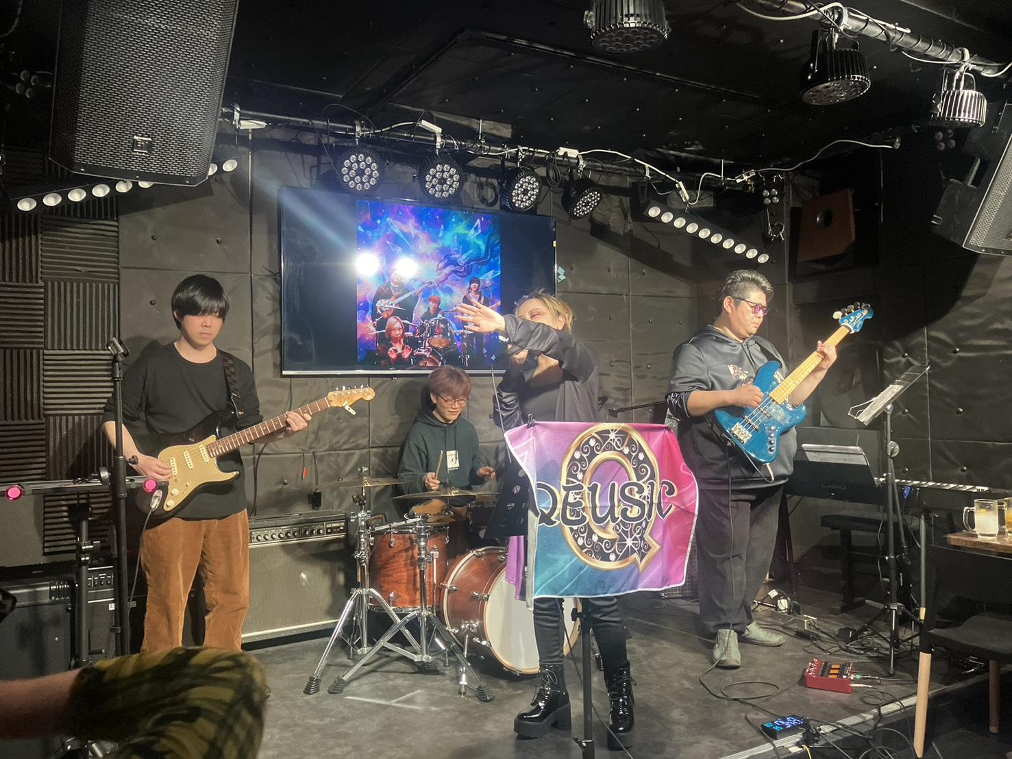 QEUSIC旗の前で歌うJUN。左にみっくん、右にのすけ、奥にまこ