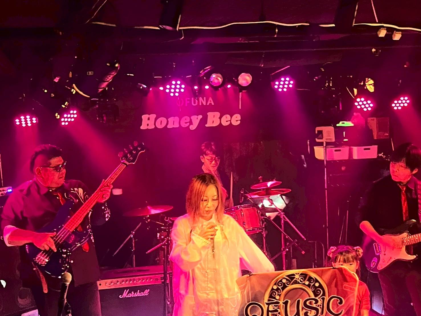マゼンタの照明に包まれた大船Honey Bee。4人がフルで演奏し、手前に子どもが旗のそばに立つ