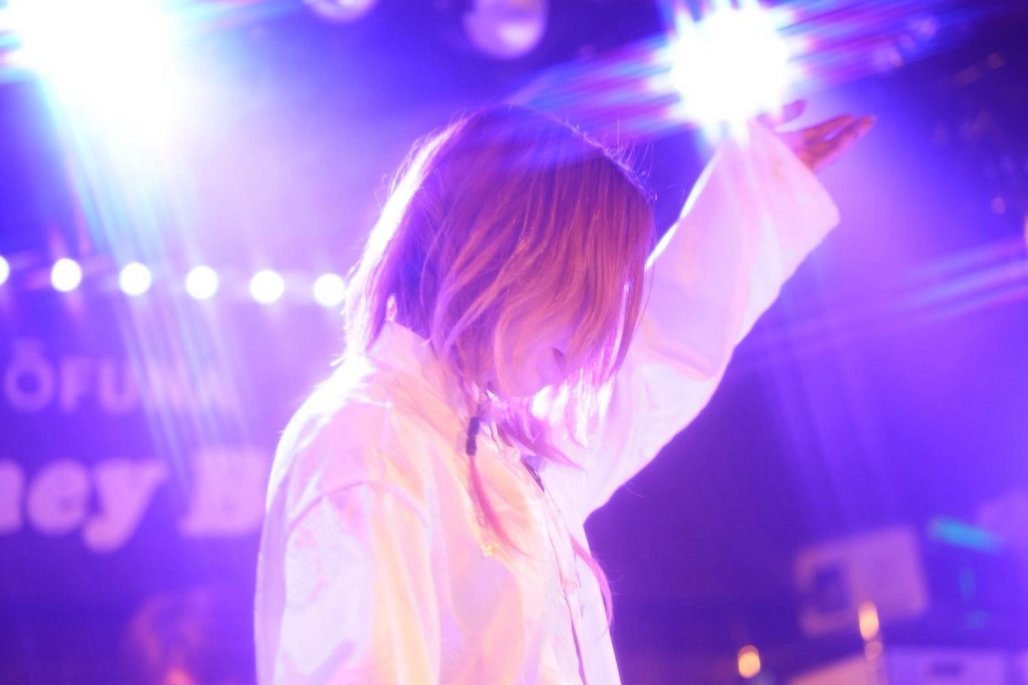 紫の照明に手を伸ばす JUN。白いジャケット、Honey Bee