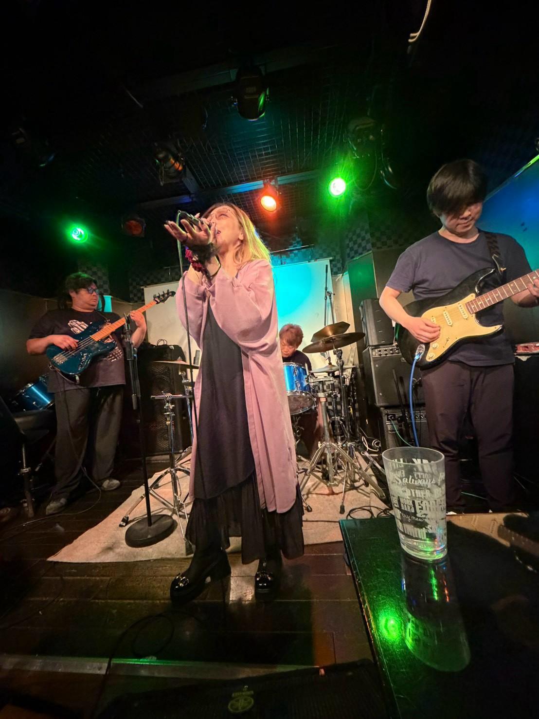 ピンクのロングカーディガンのJUNがマイクを空に掲げ、のすけとみっくんが両脇で演奏