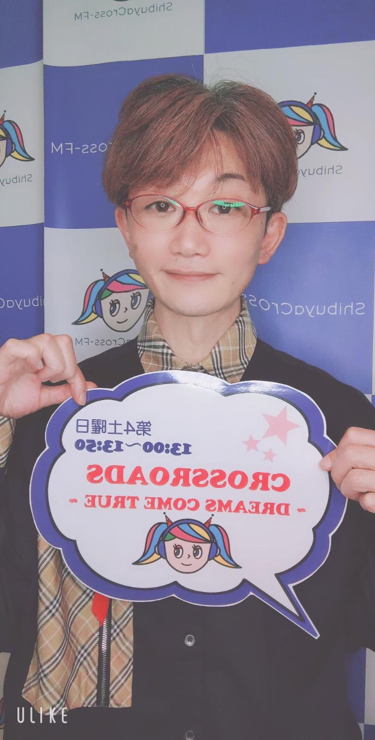 ShibuyaCross-FM、CROSSROADSの看板を持つ まこ。赤いメガネ