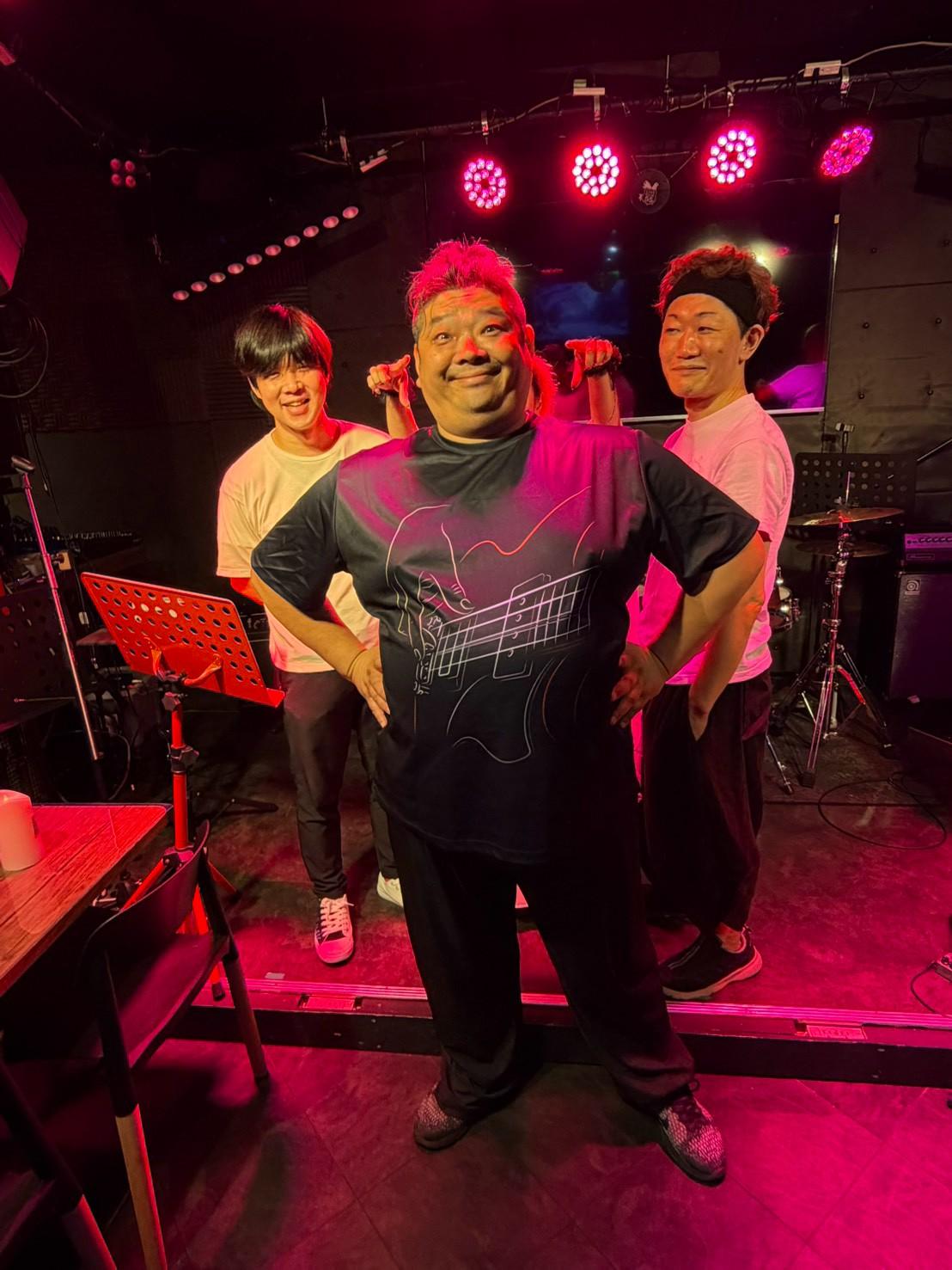 赤い照明のステージ。ベースTシャツののすけを中心に、みっくん（左）とまこ（右）が肩に手を置く