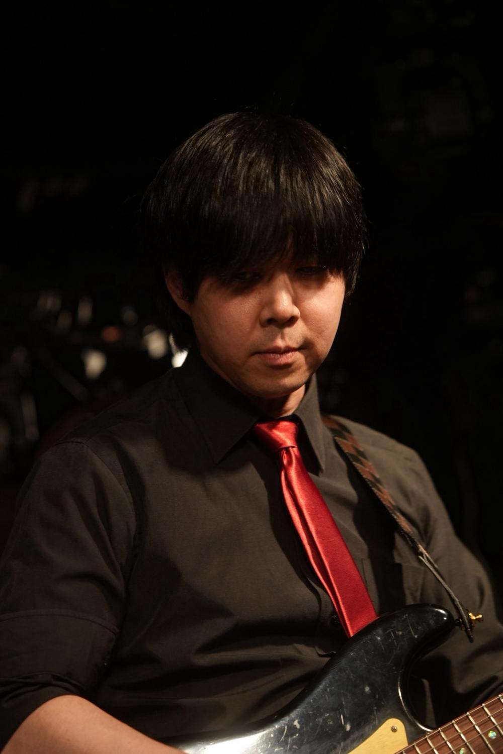 暗いステージ、赤いサテンのネクタイ、黒いストラトを構える みっくん