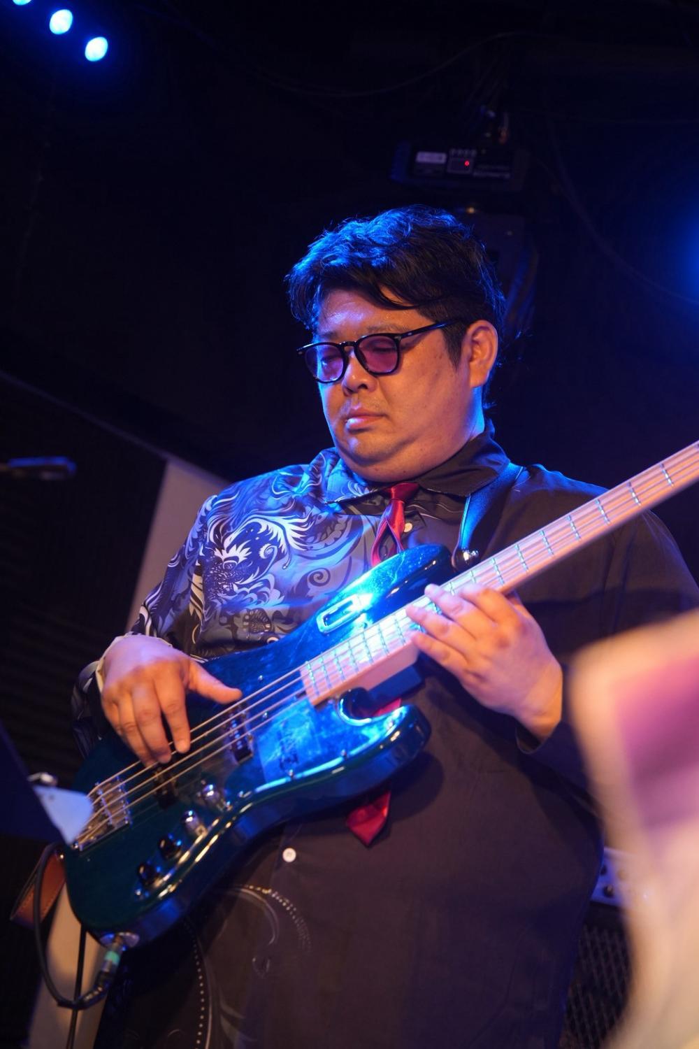 青い照明、ティールのジャズベースを構える のすけ。柄シャツ、赤いネクタイ