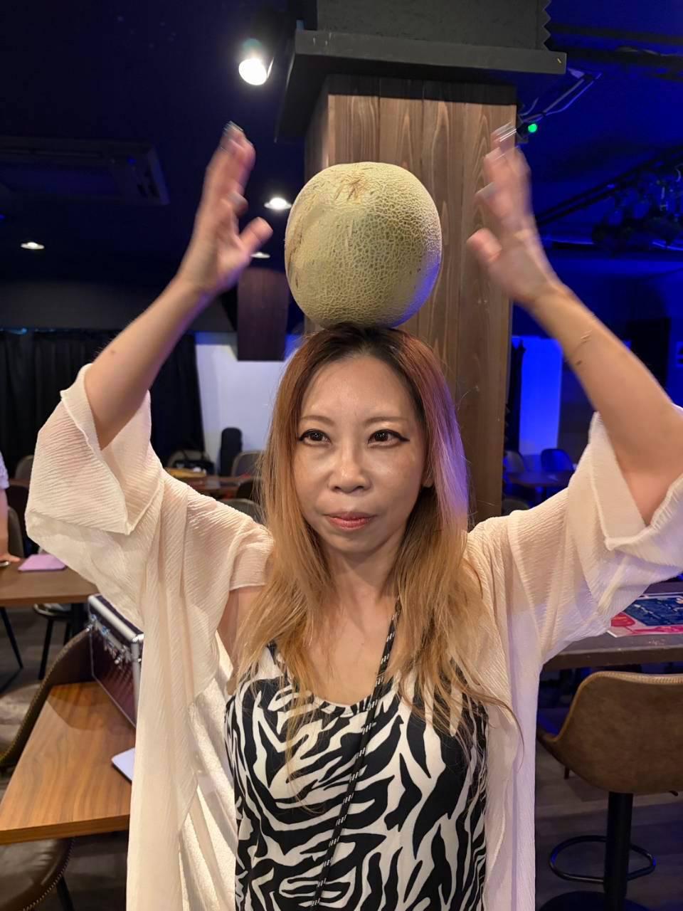 メロンを頭に乗せて両手を上げる JUN。ゼブラ柄のトップス