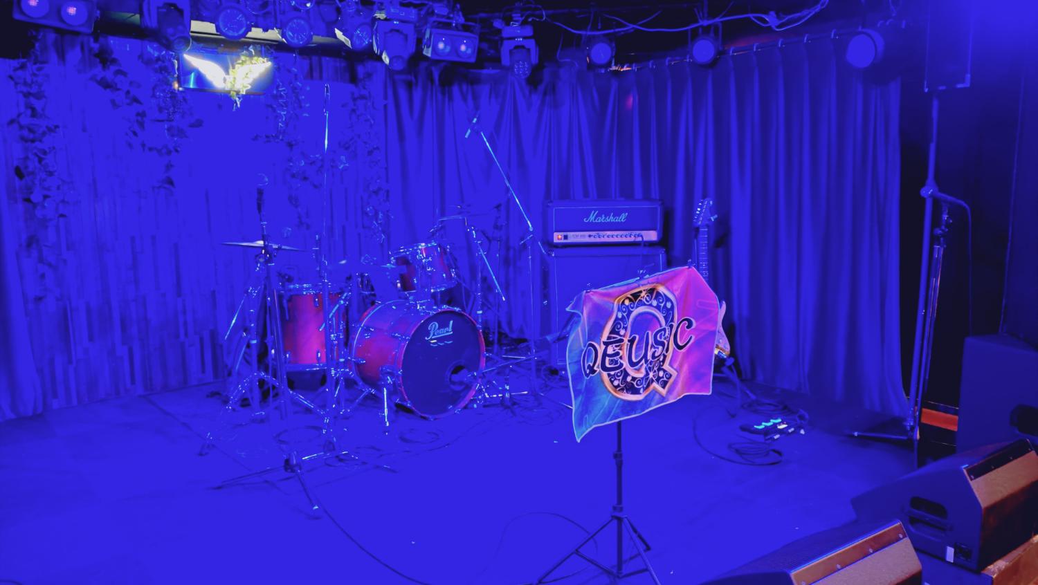 青い照明の空のステージ。QEUSIC旗、Pearlドラム、Marshallアンプ